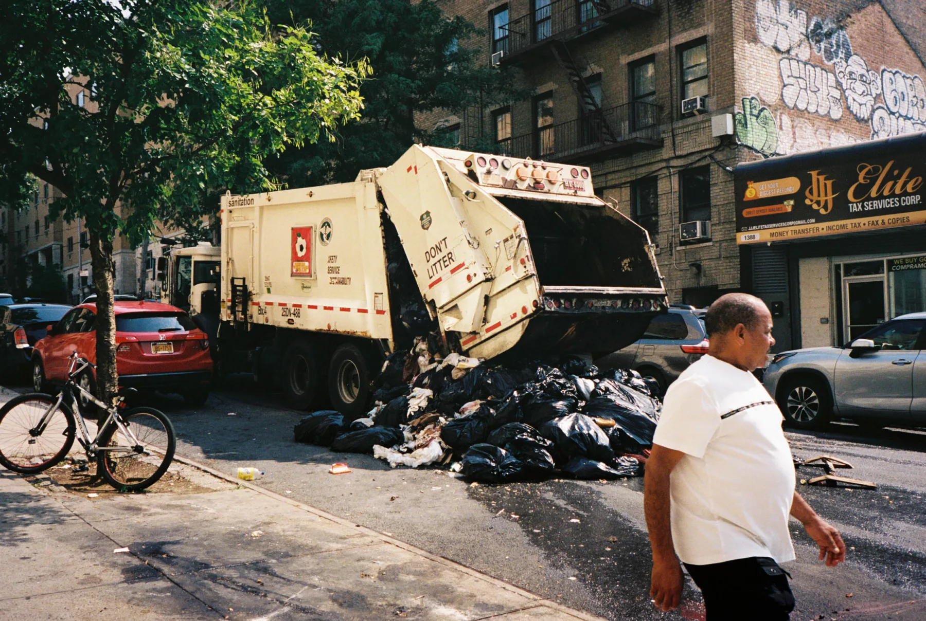 A sanitation truck with its hopper raised beside a pile of black trash bags on a Bronx street, as a man walks past.